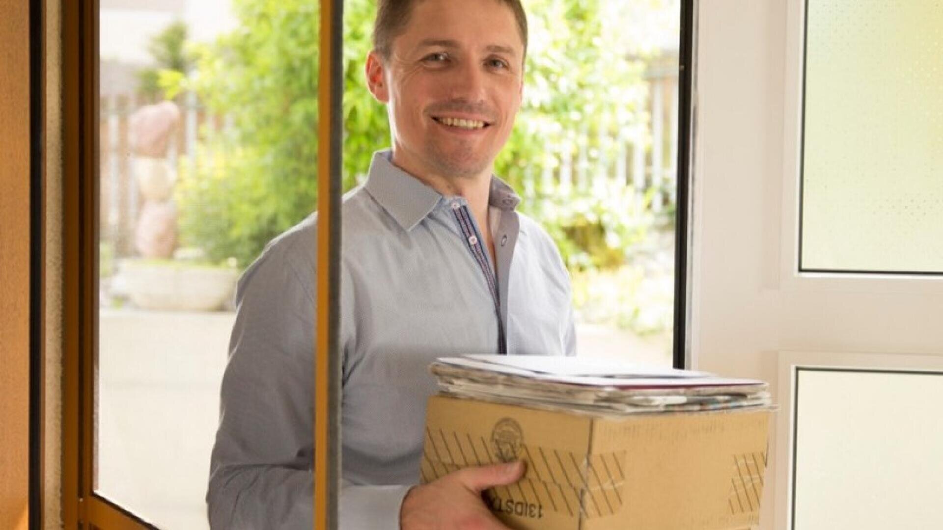 Ein lächelnder Mann steht an einer offenen Tür, in der Hand einen Karton und mehrere Briefe oder Pakete, hinter ihm ist ein heller Hintergrund im Freien zu sehen.