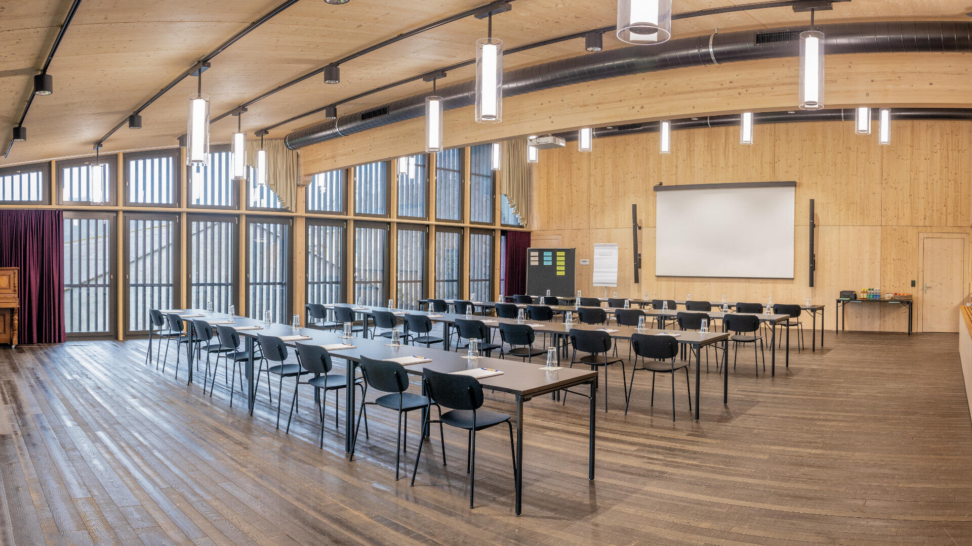 Ein geräumiger, moderner Konferenzraum mit Holzwänden und -böden, Reihen von Tischen und Stühlen vor einer großen Projektionsfläche und hohen Fenstern, die viel Tageslicht hereinlassen.