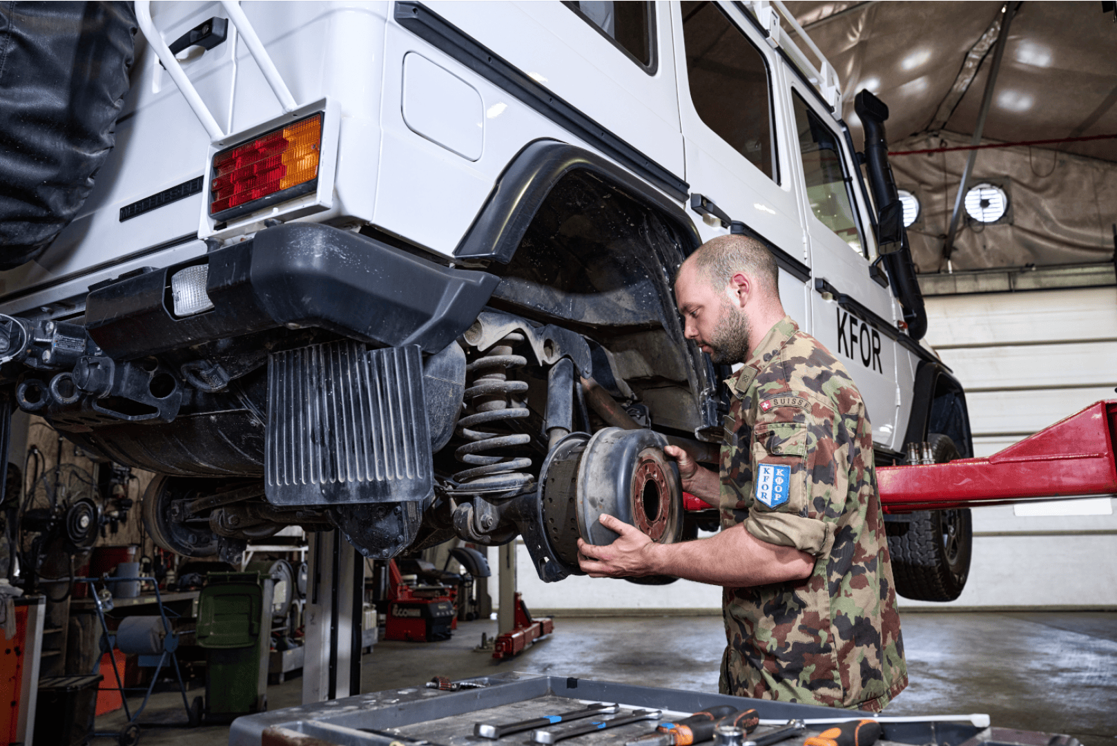 Ein Mann in Tarnuniform repariert das Hinterrad eines weißen Fahrzeugs, das auf einer Hebebühne in einer Garage angehoben wurde. Auf einer Werkbank in der Nähe liegen verschiedene Werkzeuge bereit.