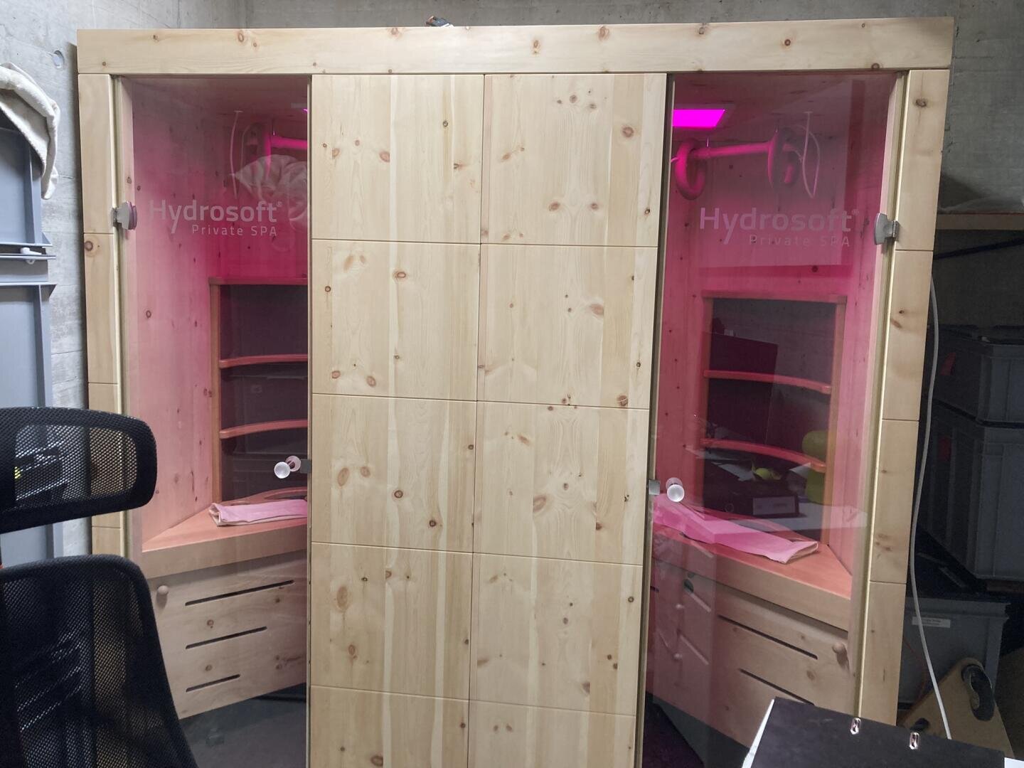 Eine hölzerne Saunakabine mit der Aufschrift Hydrosoft Private SPA mit zwei Abteilen, jedes mit Regalen, Schubladen und rosa LED-Beleuchtung im Inneren. In der Nähe befinden sich ein Bürostuhl und Ablagefächer.