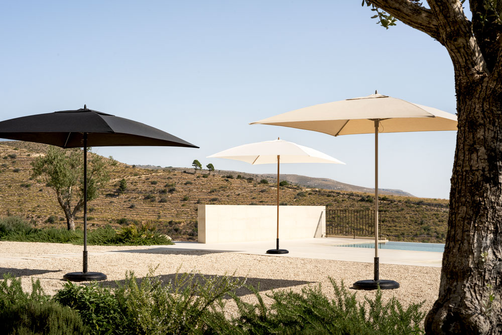 Drei grosse Sonnenschirme – ein schwarzer und zwei weisse – stehen auf einer Kiesterrasse in der Nähe eines Swimmingpools, mit einem Baum im Vordergrund und einer malerischen, hügeligen Landschaft im Hintergrund unter einem klaren Himmel.