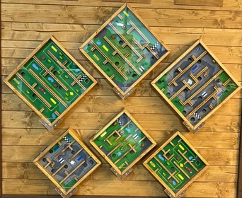 Sechs hölzerne Labyrinthe sind diagonal an einer Holzwand angebracht, jedes mit bunten Wegen, Hindernissen und kleinen Objekten, die an eine spielerische, interaktive Ausstellung erinnern.