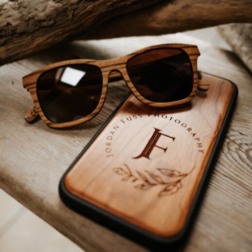 Eine hölzerne Sonnenbrille ruht auf einem hölzernen Telefonetui mit Gravur.