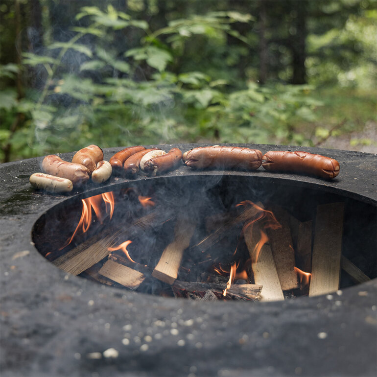 Würstchen braten auf einem flachen, runden Grill über einem Holzfeuer, aus dem Rauch aufsteigt. Grünes Laub und Bäume sind im unscharfen Hintergrund zu sehen, was auf eine Umgebung im Freien hindeutet.