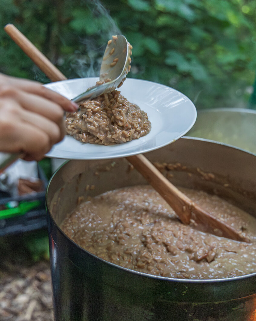 Eine Person serviert eine Portion cremig-braunes Risotto aus einem großen Topf im Freien auf einen weißen Teller, während im Hintergrund Dampf aufsteigt und grünes Laub zu sehen ist.
