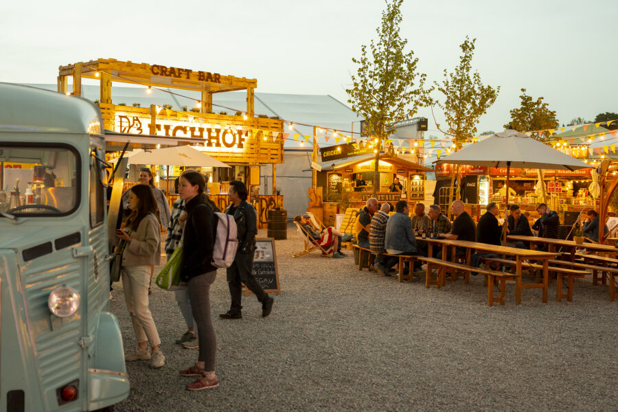 Auf einem Markt im Freien mit Lichterketten, hölzernen Essensständen, langen Picknicktischen und einem blauen Foodtruck im Vordergrund versammeln sich die Menschen und genießen Speisen und Getränke am Abend.
