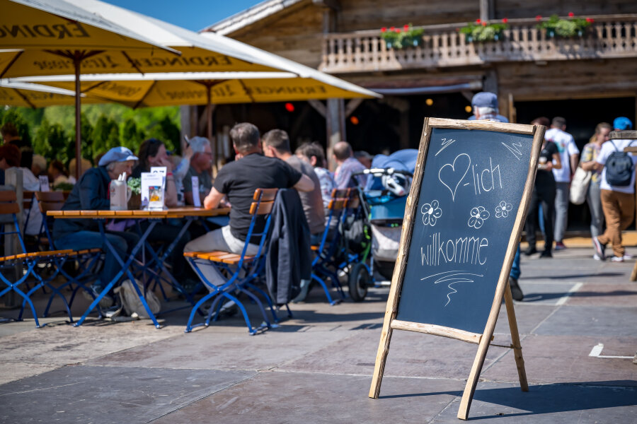Ein Kreideschild mit der Aufschrift Herzlich Willkommen steht vor einem belebten Straßencafé, in dem an einem sonnigen Tag Menschen an Tischen unter großen Sonnenschirmen sitzen. Im Hintergrund ist ein rustikales Holzgebäude zu sehen.