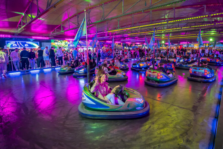 Menschen fahren Autoscooter unter bunten Neonlichtern in einem überfüllten Vergnügungspark. Im Vordergrund winkt eine Person und lächelt in die Kamera, während andere fahren oder hinter einer Absperrung zusehen. Die Atmosphäre ist lebhaft und festlich.