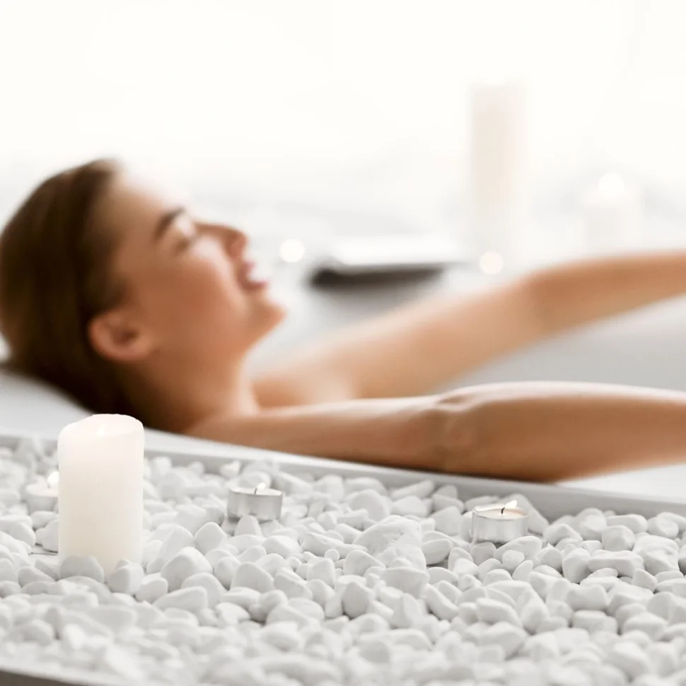 Im Hintergrund entspannt sich eine Frau in einer Badewanne, im Vordergrund liegen weiße Kieselsteine und mehrere brennende Kerzen am Wannenrand.