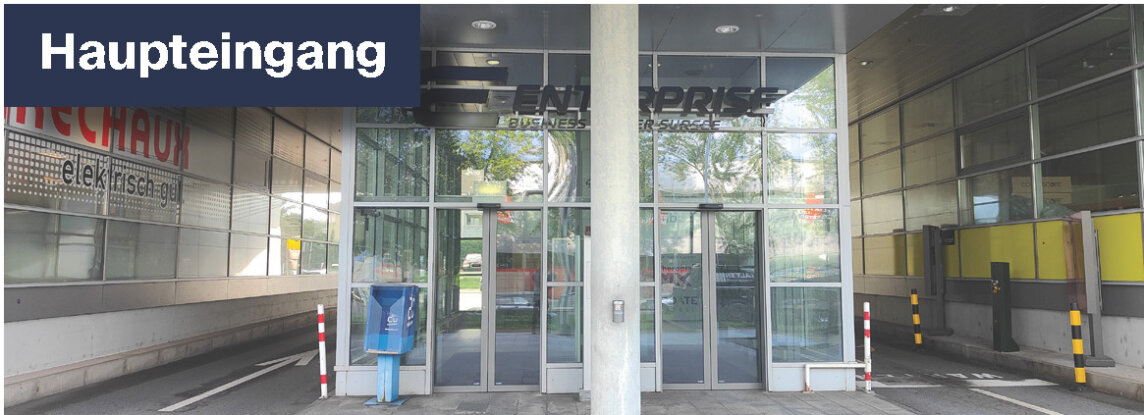 Gläserner Haupteingang eines modernen Gebäudes mit automatischen Türen, einem blauen Zeitungsständer und einem großen Schild mit der Aufschrift "Haupteingang" darüber. Der Bereich ist teilweise überdacht und wird von Glaswänden flankiert.