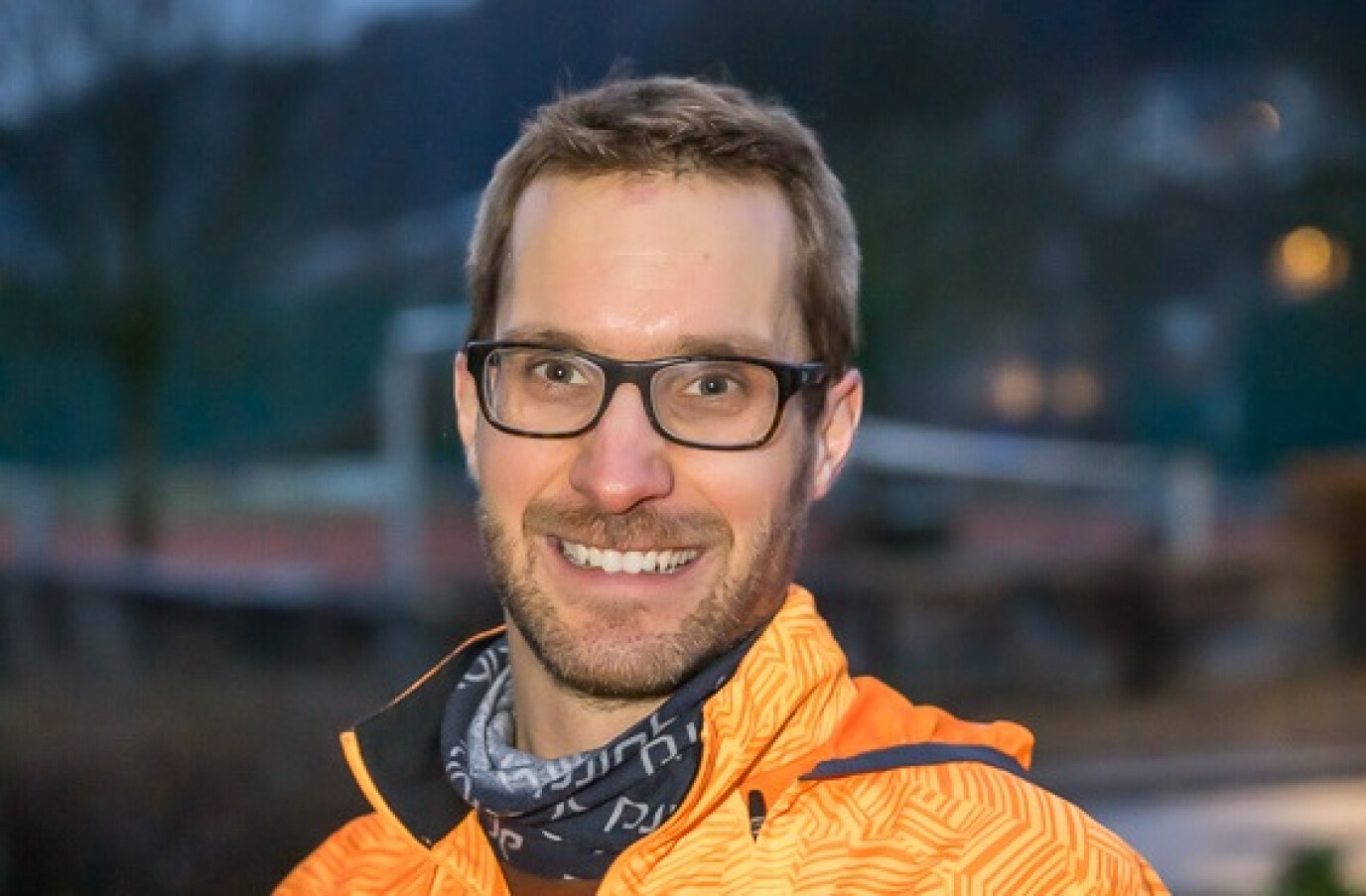 Ein Mann mit Brille und kurzem braunem Haar trägt eine auffällig orangefarbene Laufjacke und einen schwarzen Schal. Er lächelt und steht auf einer Sportanlage in der Abenddämmerung.