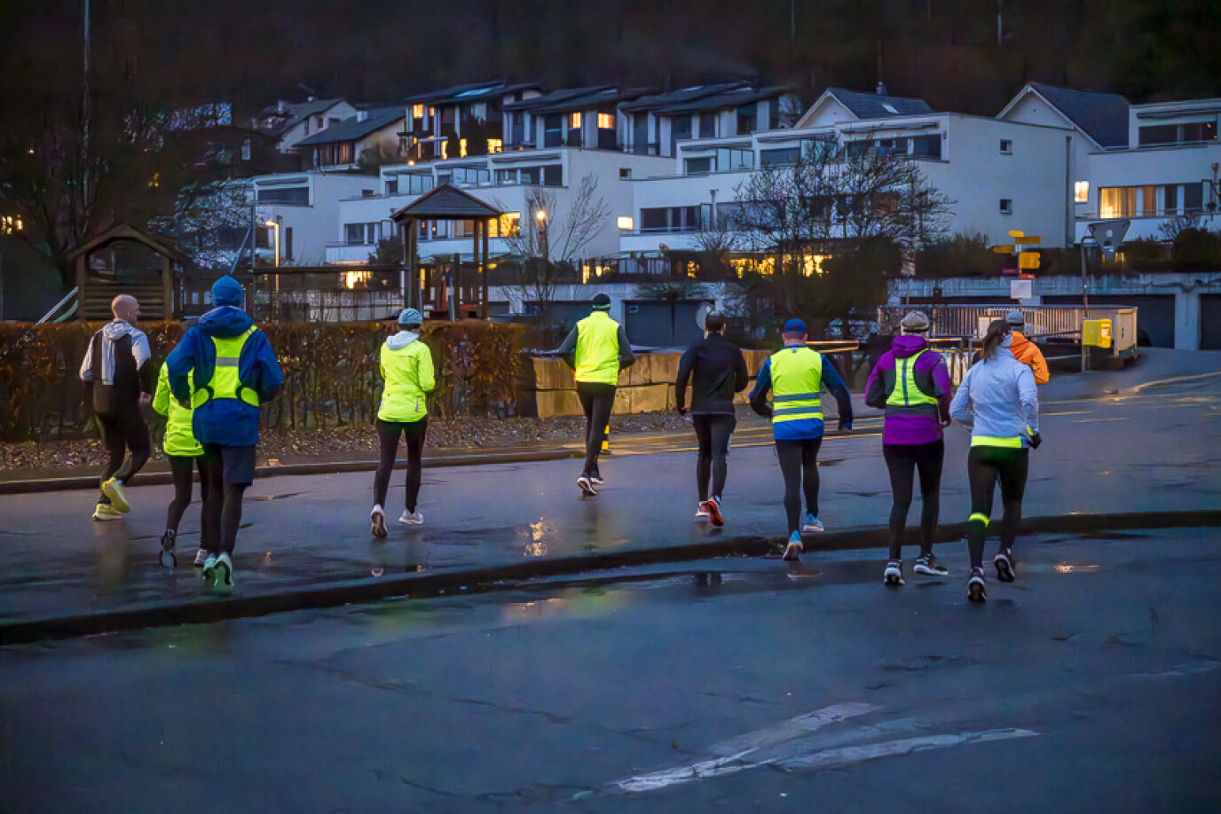 Eine Gruppe von Joggerinnen und Joggern läuft in der Dämmerung auf nasser Straße durch ein Wohnquartier. Die meisten tragen reflektierende oder leuchtende Sportkleidung. Im Hintergrund sind beleuchtete Wohnhäuser zu sehen, die eine gemütliche Abendstimmung vermitteln.