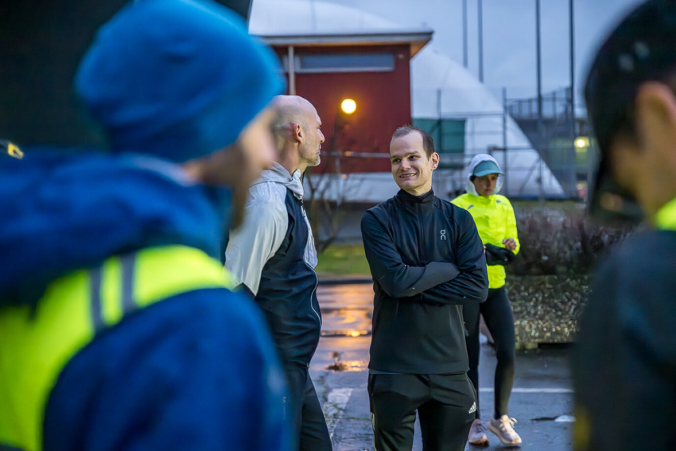 Ein junger Mann in schwarzer Sportkleidung steht mit verschränkten Armen und lächelt im Gespräch mit einem anderen Jogger. Um sie herum stehen weitere Personen in heller Sportkleidung. Die Szene spielt sich auf einem Sportplatz bei Dämmerung ab.