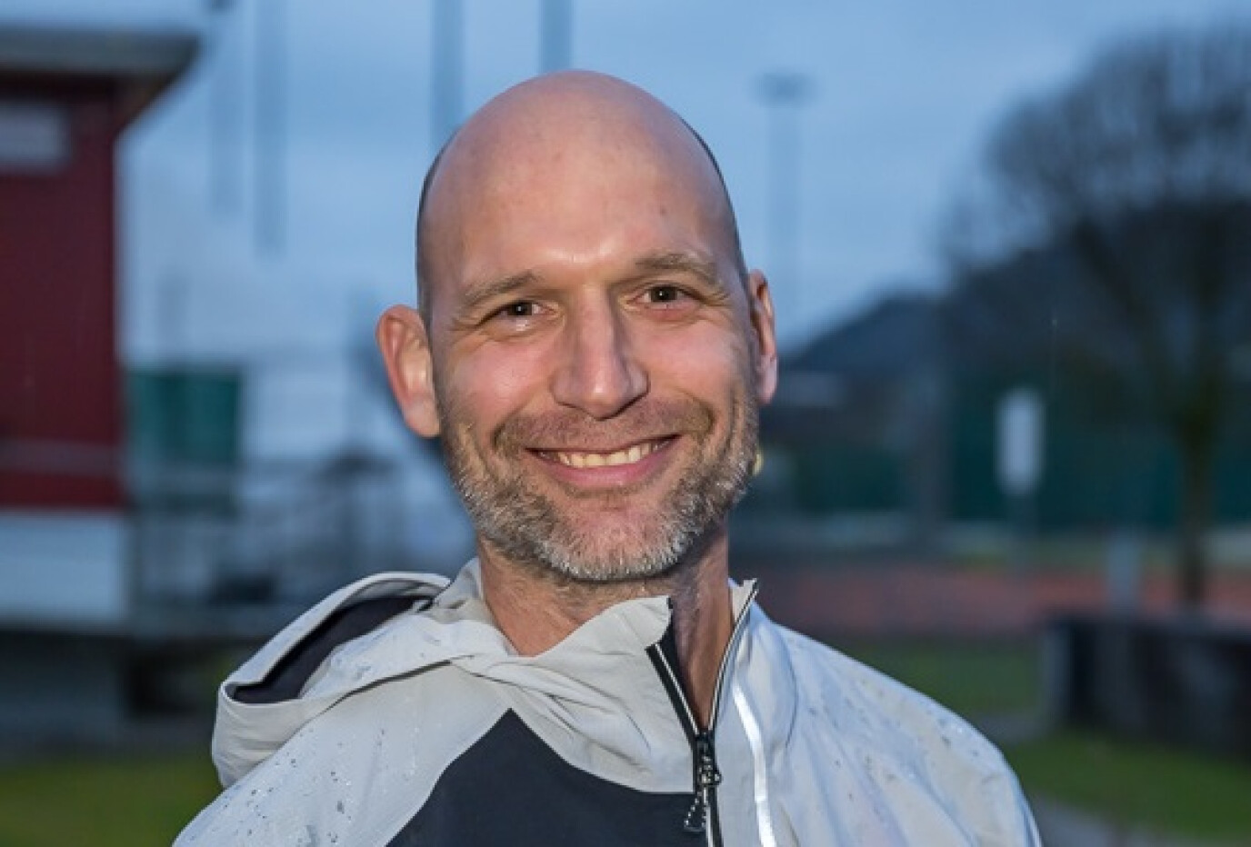 Ein Mann mit Glatze und kurzem Bart, der ein weiss-schwarzes Sportoutfit trägt, lächelt in die Kamera. Im Hintergrund ist eine Sportanlage bei Dämmerung sichtbar.