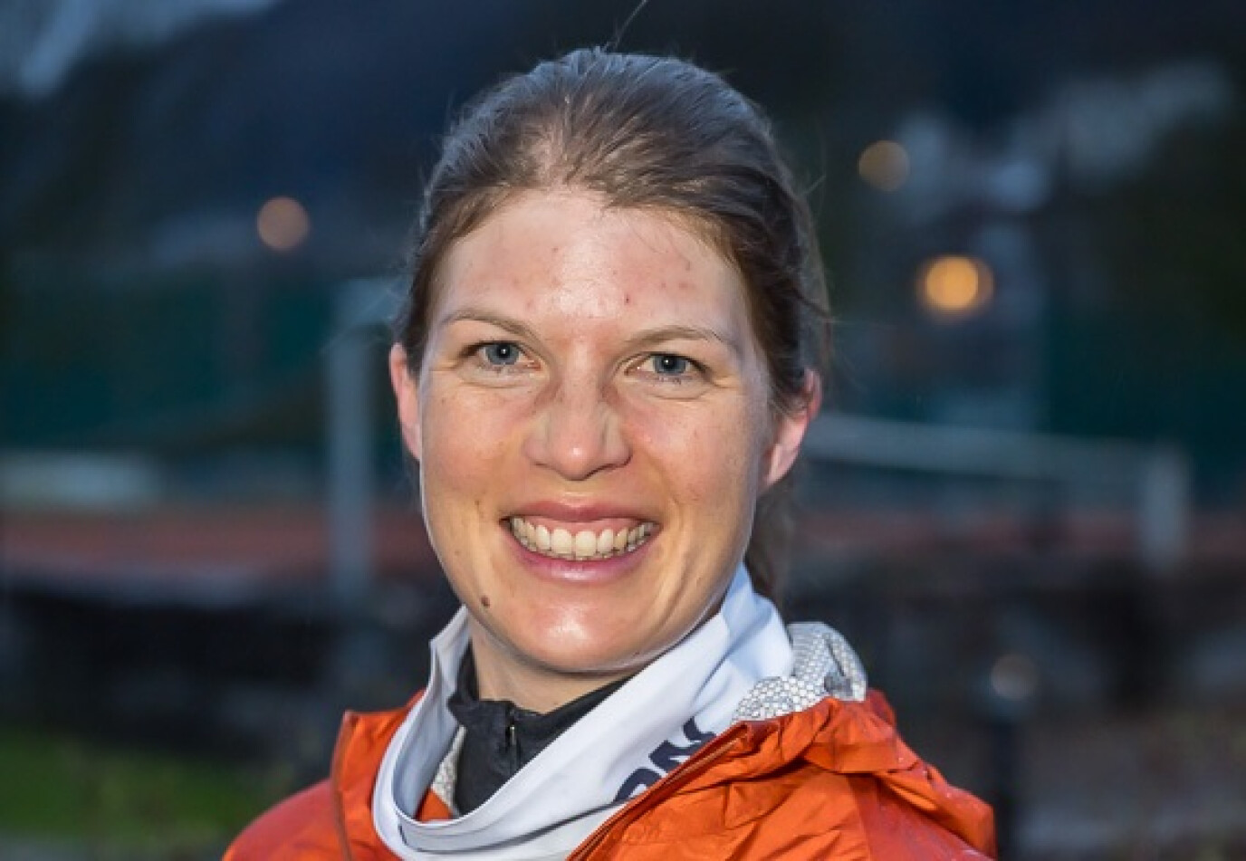 Eine Frau mit braunen Haaren, die zu einem Zopf gebunden sind, trägt eine orangefarbene Jacke und einen weißen Schal. Sie lächelt in die Kamera, während sich der Hintergrund unscharf zeigt.