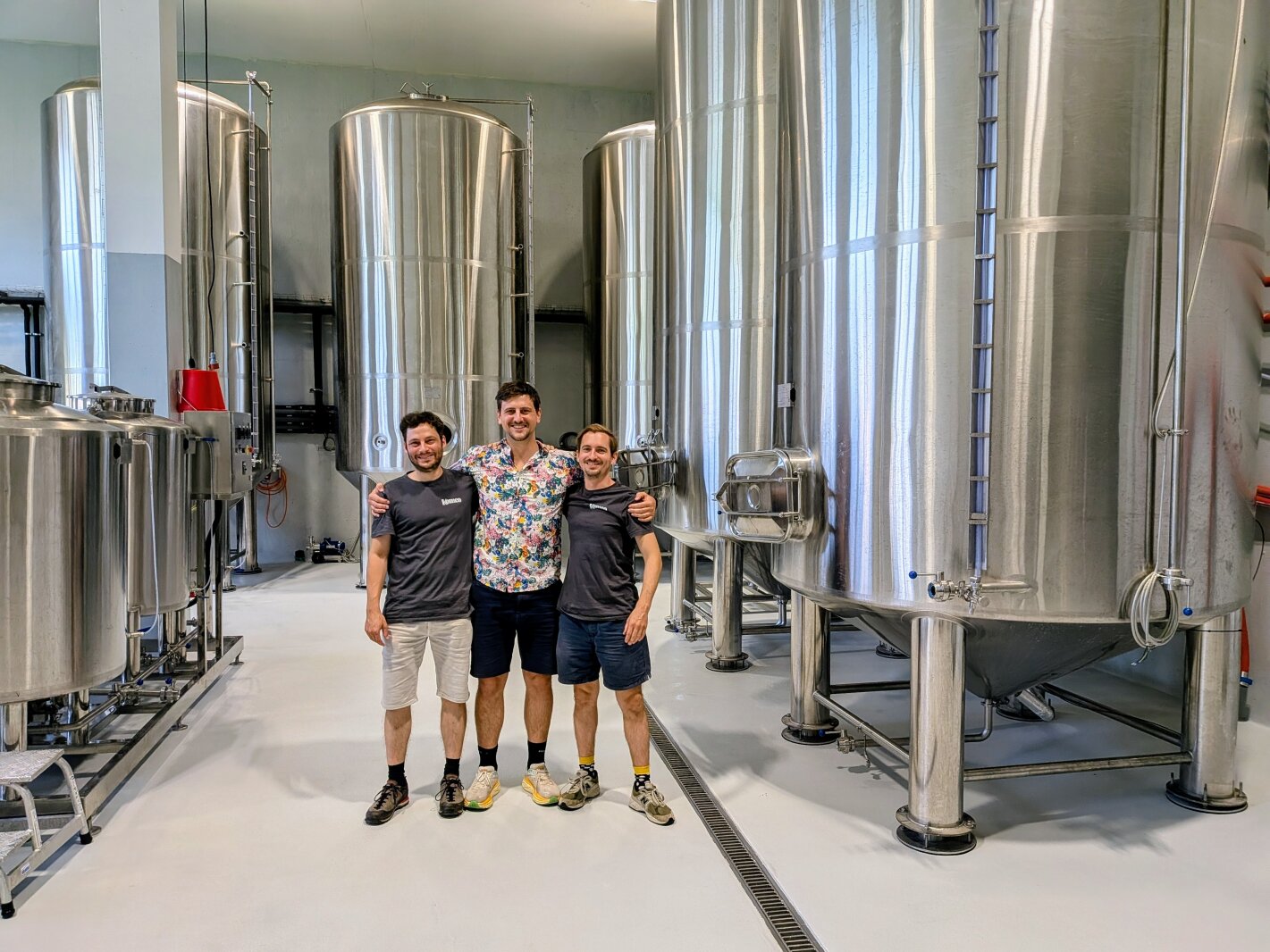Drei Personen stehen lächelnd mit umschlungenen Armen in einer Brauerei, umgeben von großen Gärtanks und Brauanlagen aus rostfreiem Stahl.