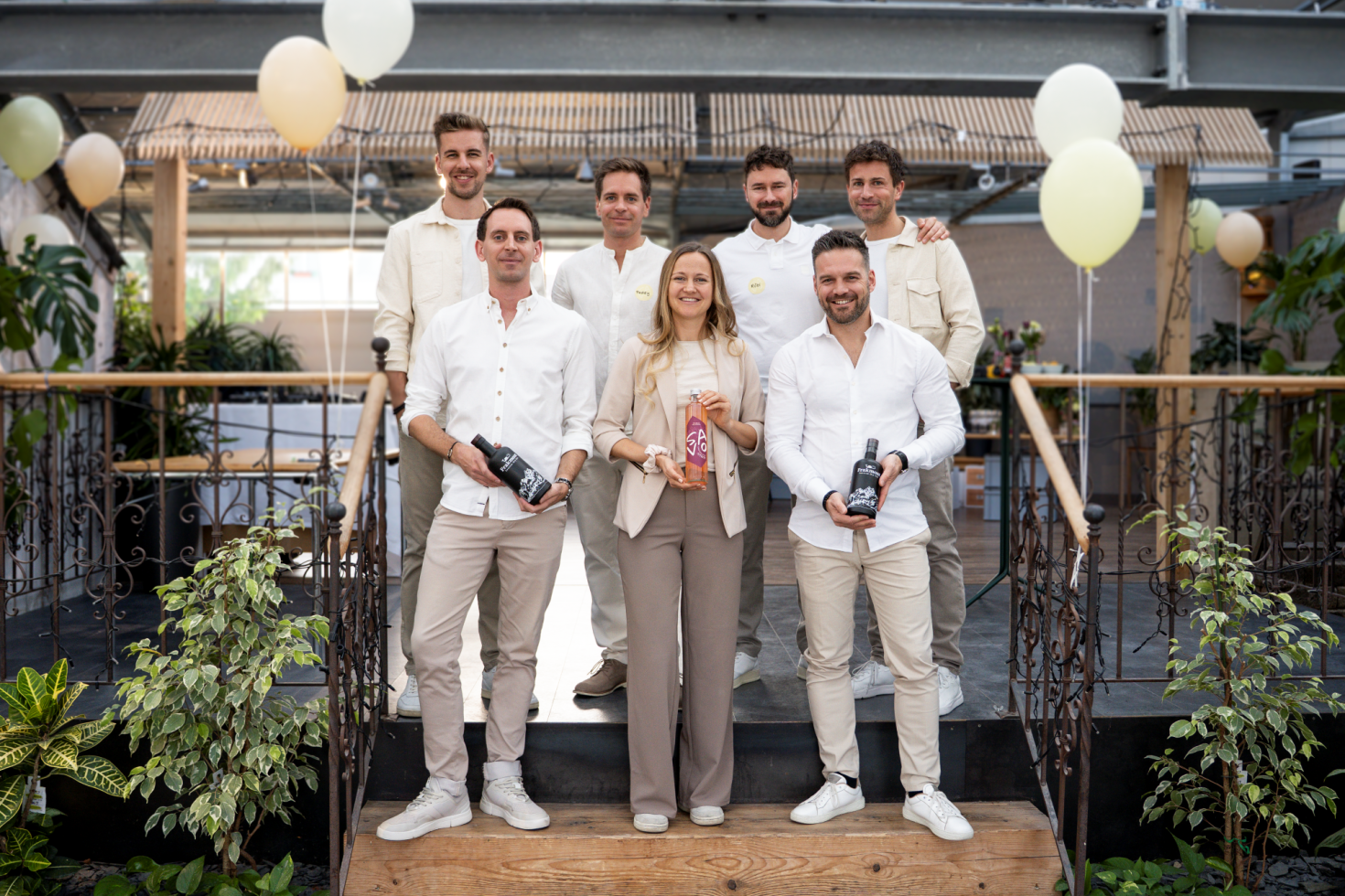 Sieben Personen in heller Kleidung stehen auf Stufen in einem dekorierten Innenraum und lächeln in die Kamera. Einige halten Flaschen oder eine Auszeichnung in der Hand. Um sie herum sind Luftballons, Grünpflanzen und Holzgeländer zu sehen.