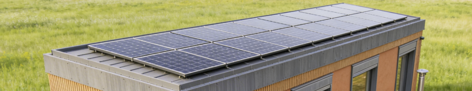 Eine Reihe von Solarmodulen ist auf dem Flachdach eines modernen kleinen Hauses mit Holzverkleidung installiert, das von grünem Gras umgeben ist.