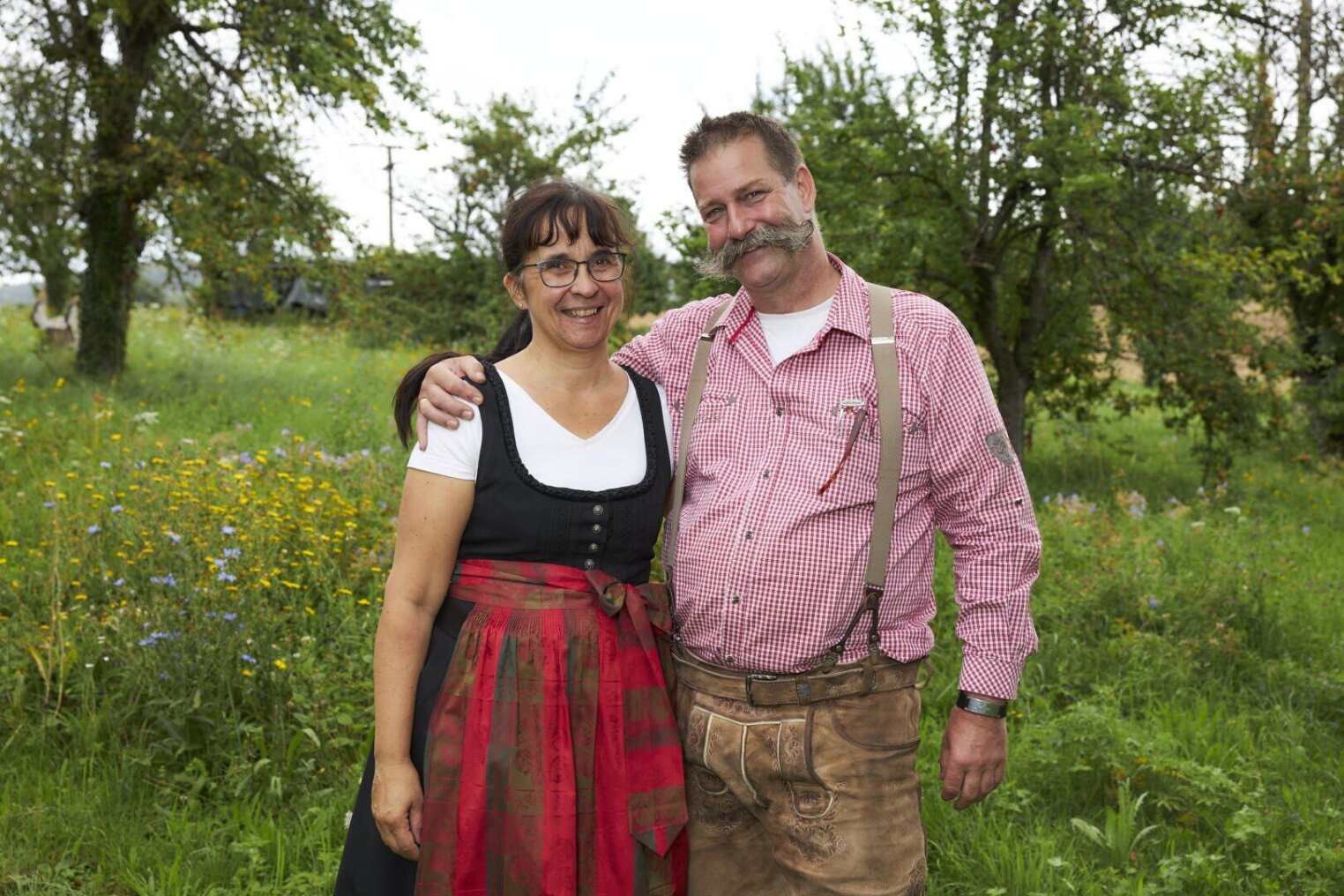 Ein lächelndes Paar steht in traditioneller bayerischer Kleidung im Freien; die Frau trägt ein Dirndlkleid, während der Mann eine Lederhose und ein kariertes Hemd trägt und seinen Arm um sie legt. Im Hintergrund sind grüne Bäume und Wildblumen zu sehen.