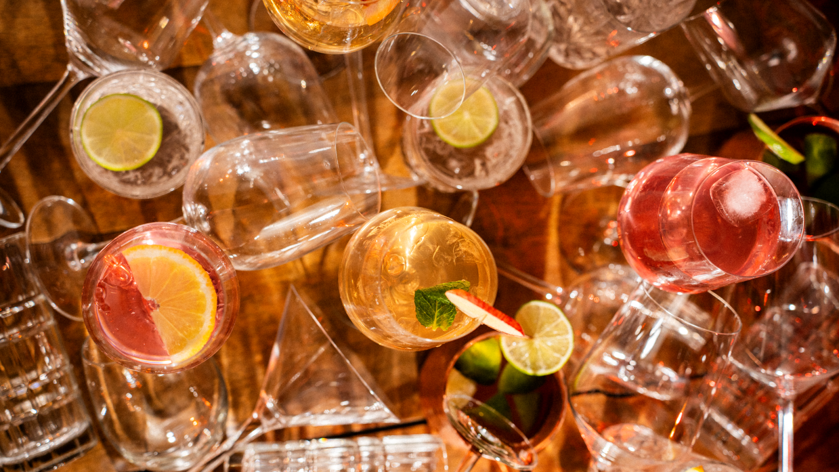 Eine Vielzahl bunter Cocktails mit Zitronen-, Limetten- und Minzgarnituren sind zwischen leeren Gläsern auf einer hölzernen Oberfläche angeordnet, von oben betrachtet.