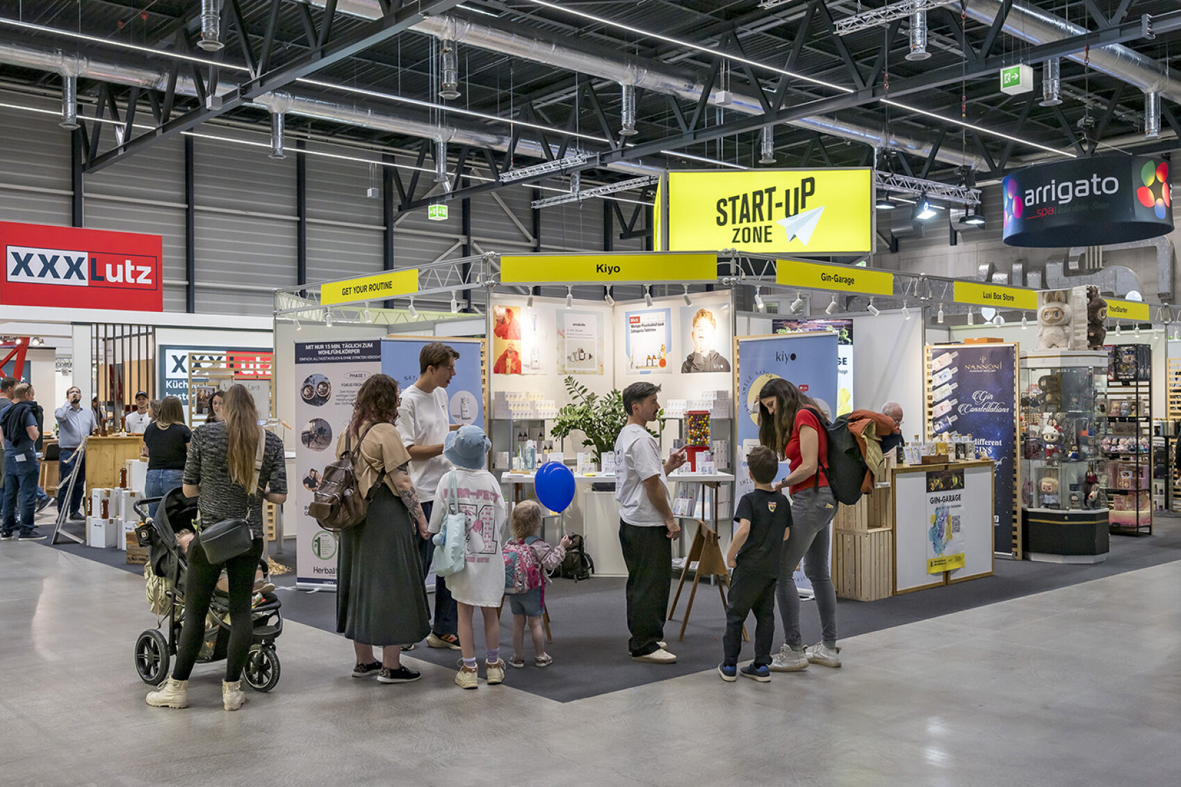 Mehrere Besuchende, darunter Familien mit Kindern, informieren sich an den Ständen der Start-up-Zone. Die jungen Unternehmen präsentieren ihre Produkte und Ideen im persönlichen Gespräch. Die offene Standgestaltung lädt zum Entdecken und Austauschen ein.