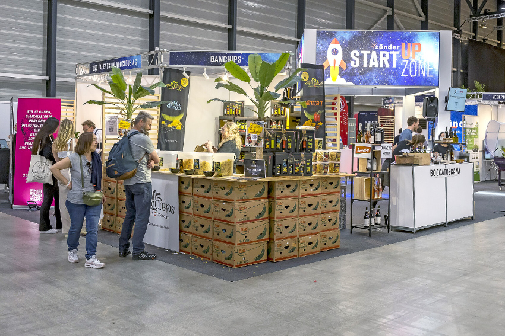 Messestand in der "Start-up Zone" mit einem Stand für Bananenchips und anderen Produkten. Mehrere Besuchende schauen sich das Sortiment an.