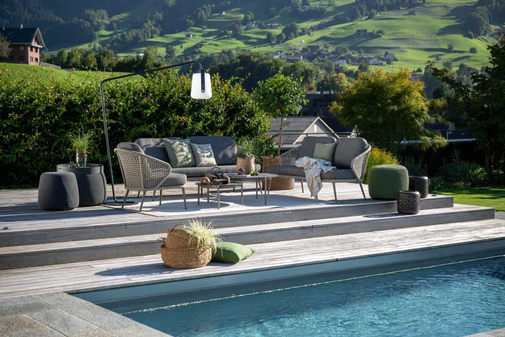 Moderne Außenterrasse mit Korbmöbeln, Kissen, einem Couchtisch und dekorativen Pflanzen auf einem Holzdeck neben einem Pool. Im Hintergrund sind malerische grüne Hügel und Häuser zu sehen.