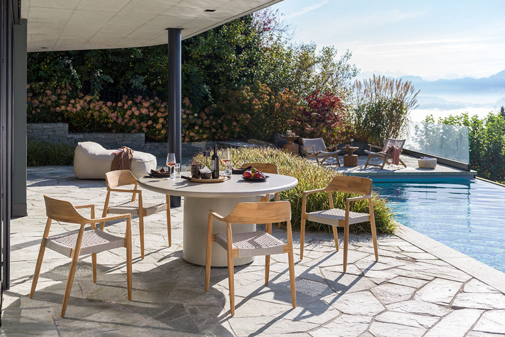 Eine moderne Außenterrasse mit einem runden Esstisch für eine Mahlzeit, umgeben von vier Holzstühlen. In der Nähe gibt es einen Pool, Liegestühle, üppiges Grün und eine malerische Aussicht im Hintergrund.