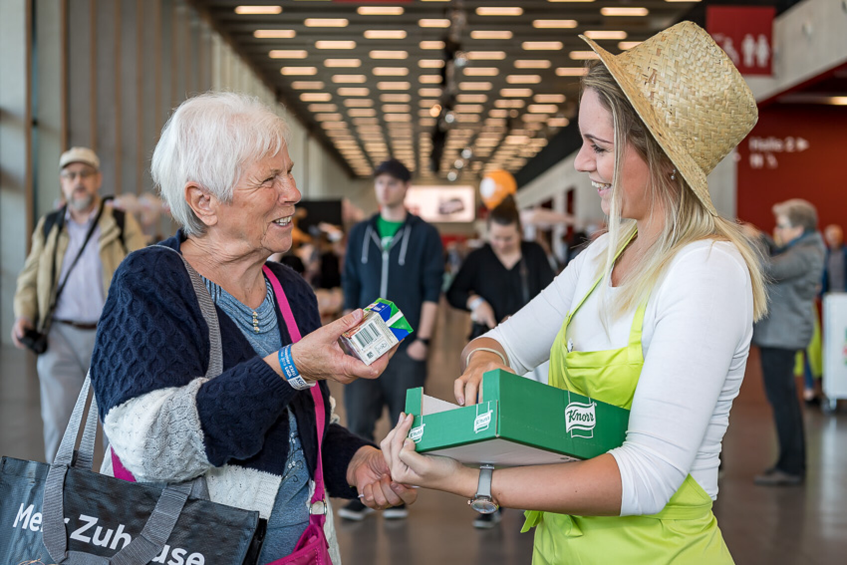 Eine Frau mit Strohhut und grüner Schürze überreicht einer älteren Dame ein Produkt aus einem grünen Karton mit der Aufschrift 'Knorr'. Beide lächeln, und im Hintergrund sind weitere Messebesucher zu sehen.