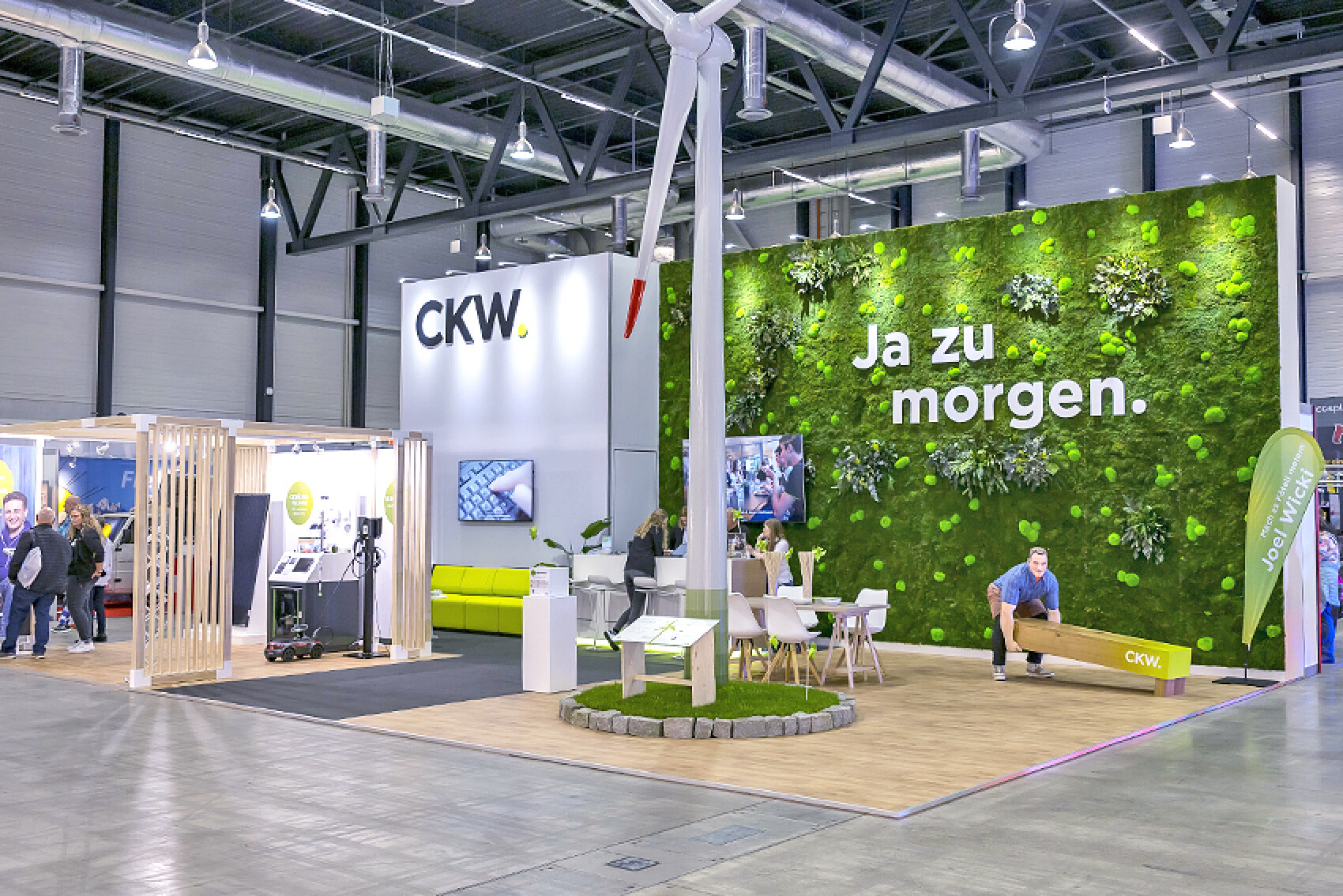 Messestand von CKW mit einer grünen Mooswand, auf der "Ja zu morgen." steht. Im Vordergrund eine Windturbine und eine Sitzgruppe, im Hintergrund Besuchende in der Messehalle.