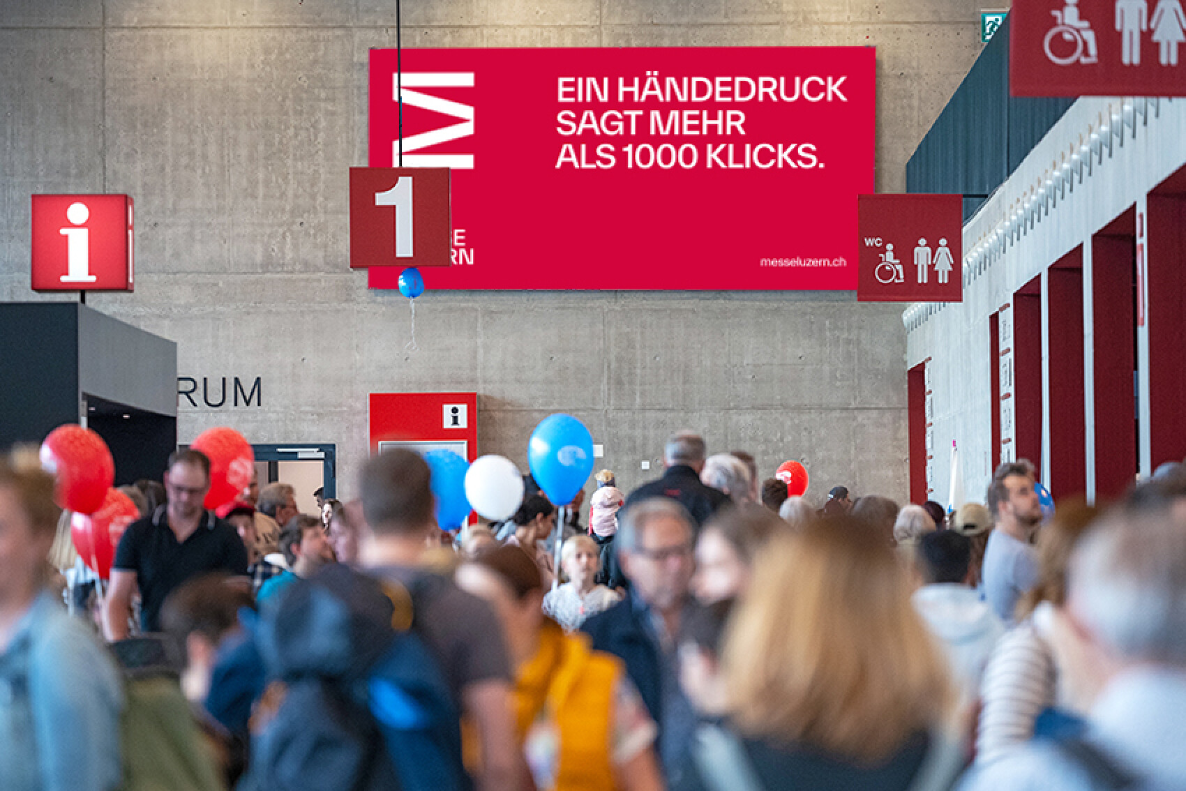 Ein grosses rotes Banner in einer Messehalle mit der Aufschrift 'Ein Händedruck sagt mehr als 1000 Klicks'. Darunter sind zahlreiche Messebesucher mit bunten Luftballons zu sehen.