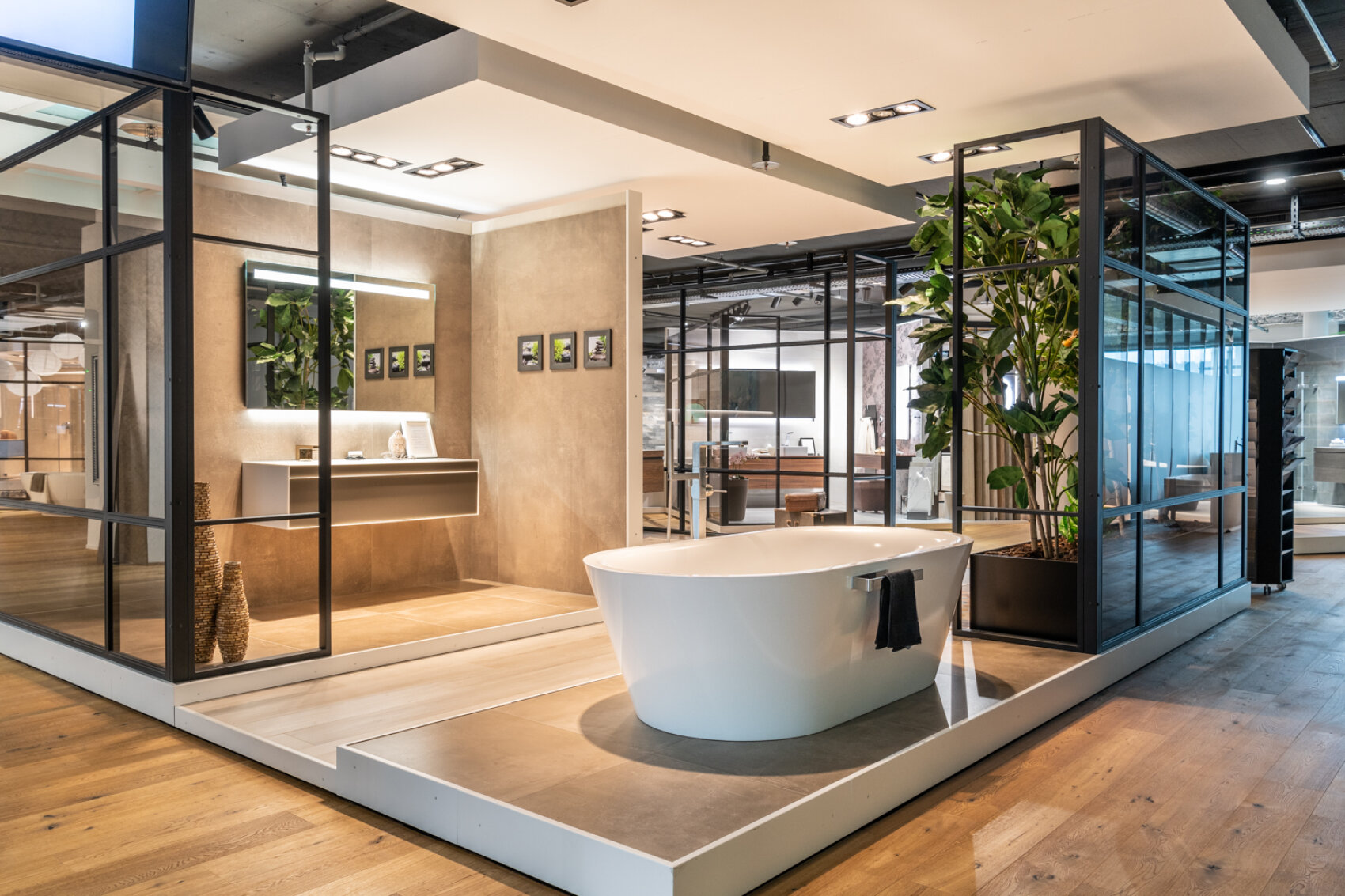 Ein moderner Ausstellungsraum mit einer freistehenden weißen Badewanne, Glaswänden, großen Pflanzen, Holzfußböden und einem eleganten Waschtisch mit Spiegel und gerahmten Fotos schafft eine stilvolle und offene Badausstellung.