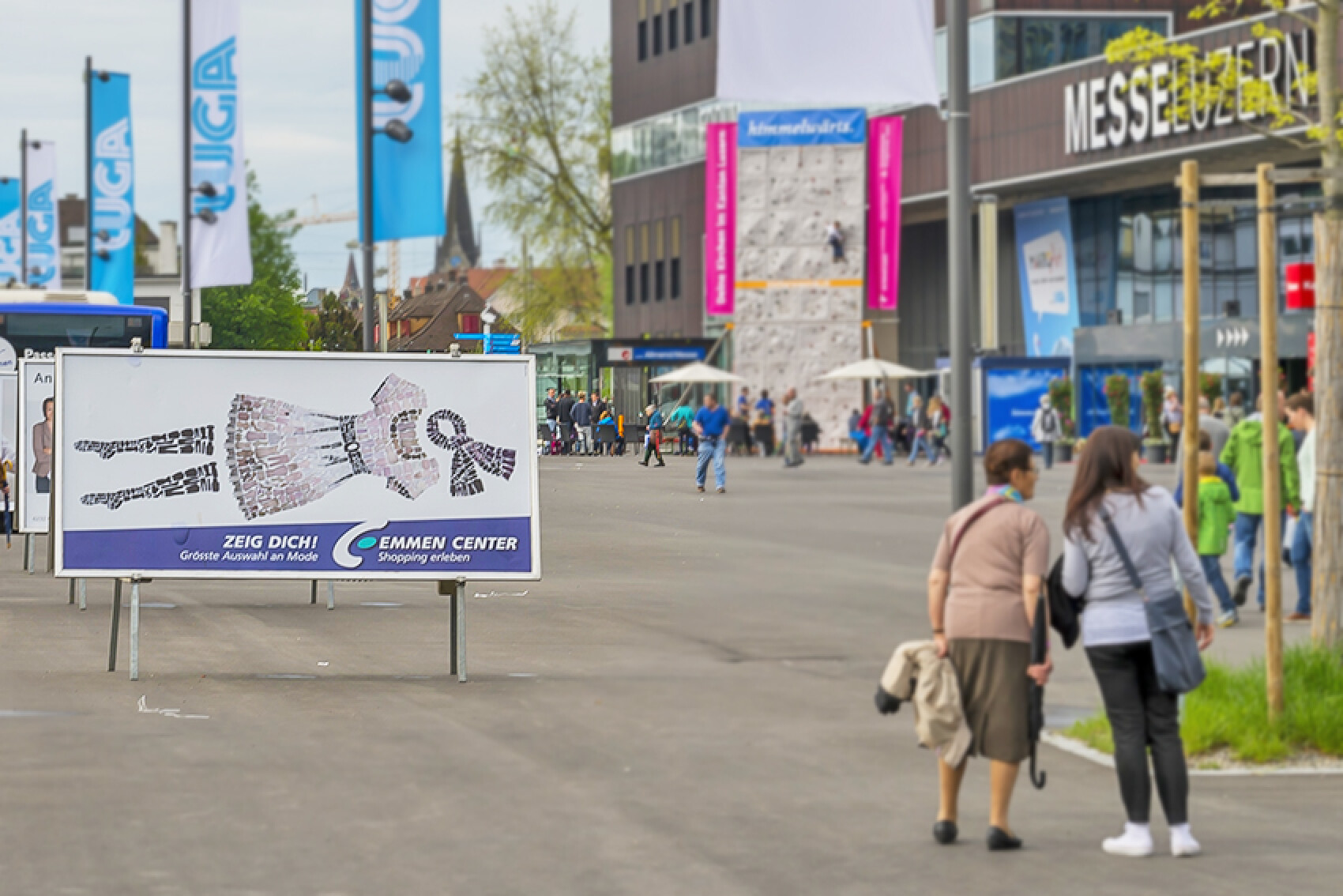 Ein grossformatiges Plakat vor der Messe Luzern mit der Aufschrift 'Zeig dich! Emmen Center – Grösste Auswahl an Mode'. Im Hintergrund sind die Messegebäude und Besucher zu sehen.