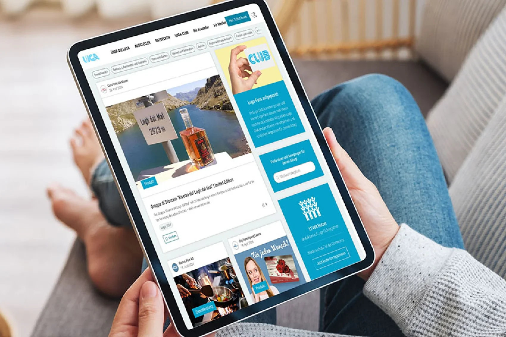 Eine Person hält ein Tablet, auf dem die Luga-365-Webseite geöffnet ist. Sie zeigt ein Bild von einem Produkt, das vor einem Bergsee präsentiert wird. Im Hintergrund sind Beine in bequemer Sitzhaltung zu sehen.