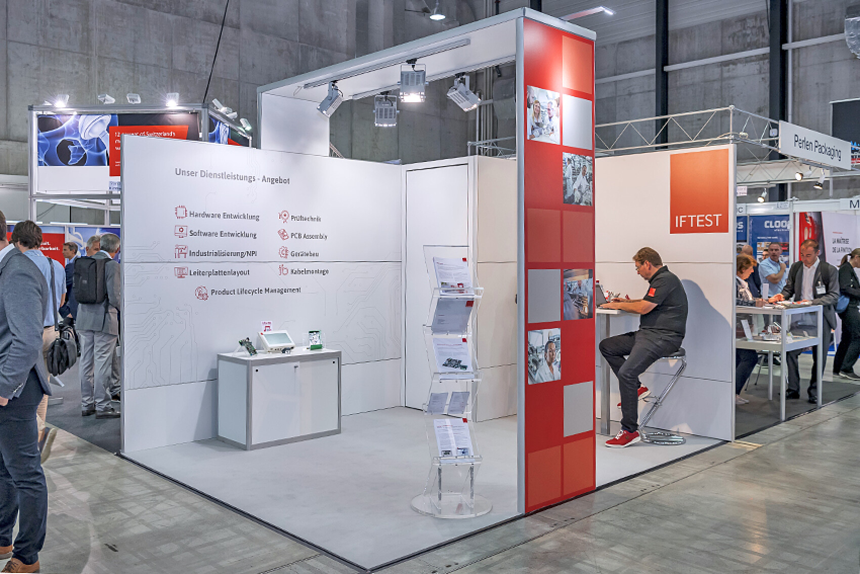 Messestand der Firma Iftest mit einer modernen Gestaltung in Weiß und Rot. Ein Mann sitzt an einer Theke und arbeitet, während sich Messebesuchende umsehen.