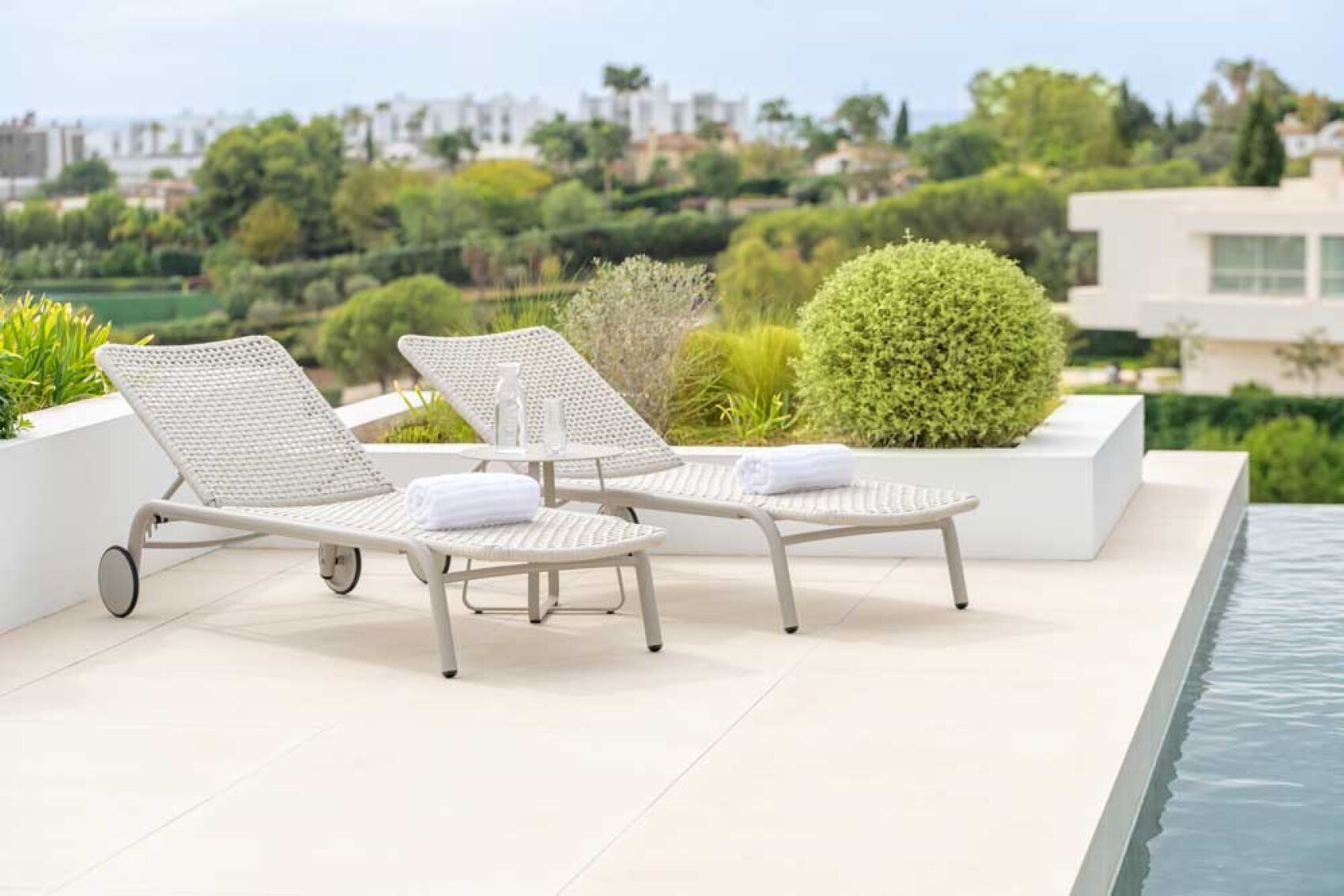 Zwei weisse, geflochtene Liegestühle mit Handtüchern und einer Wasserflasche stehen am Rande eines modernen Pools, umgeben von Grün und mit Blick auf eine malerische, gehobene Wohngegend.