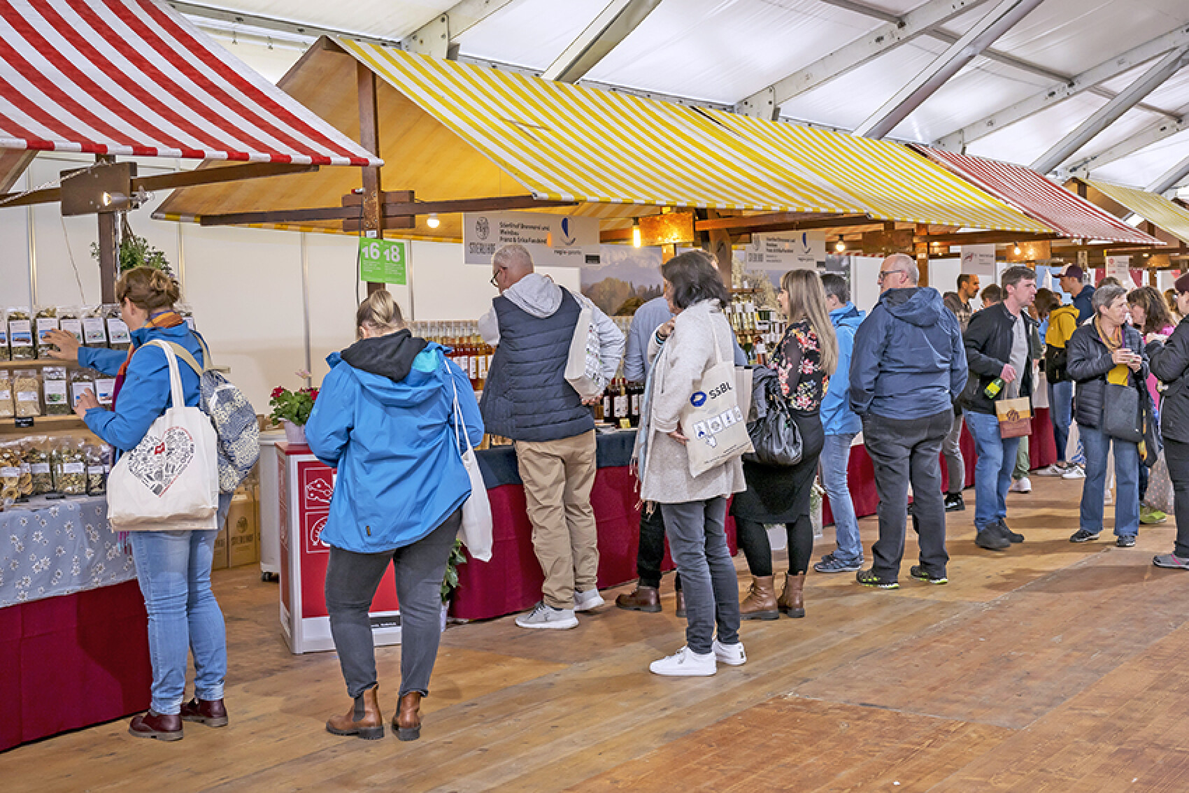 Marktstand in einer Halle mit Holzbuden und bunten Markisen. Menschen kaufen regionale Produkte ein, ein großer blauer Aufkleber mit "Ausgebucht" ist im Vordergrund.