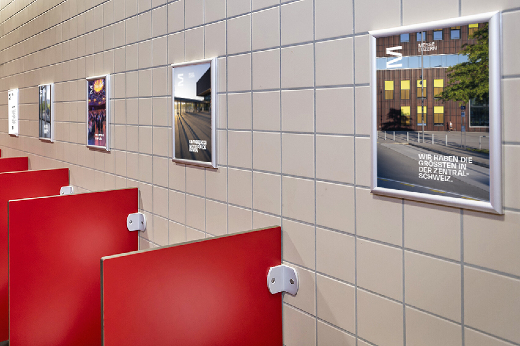 Eine Kachelwand in einer öffentlichen Toilette, an der mehrere Poster angebracht sind. Jedes Poster zeigt unterschiedliche Werbeinhalte. Im unteren Bereich sind rote Trennwände sichtbar.