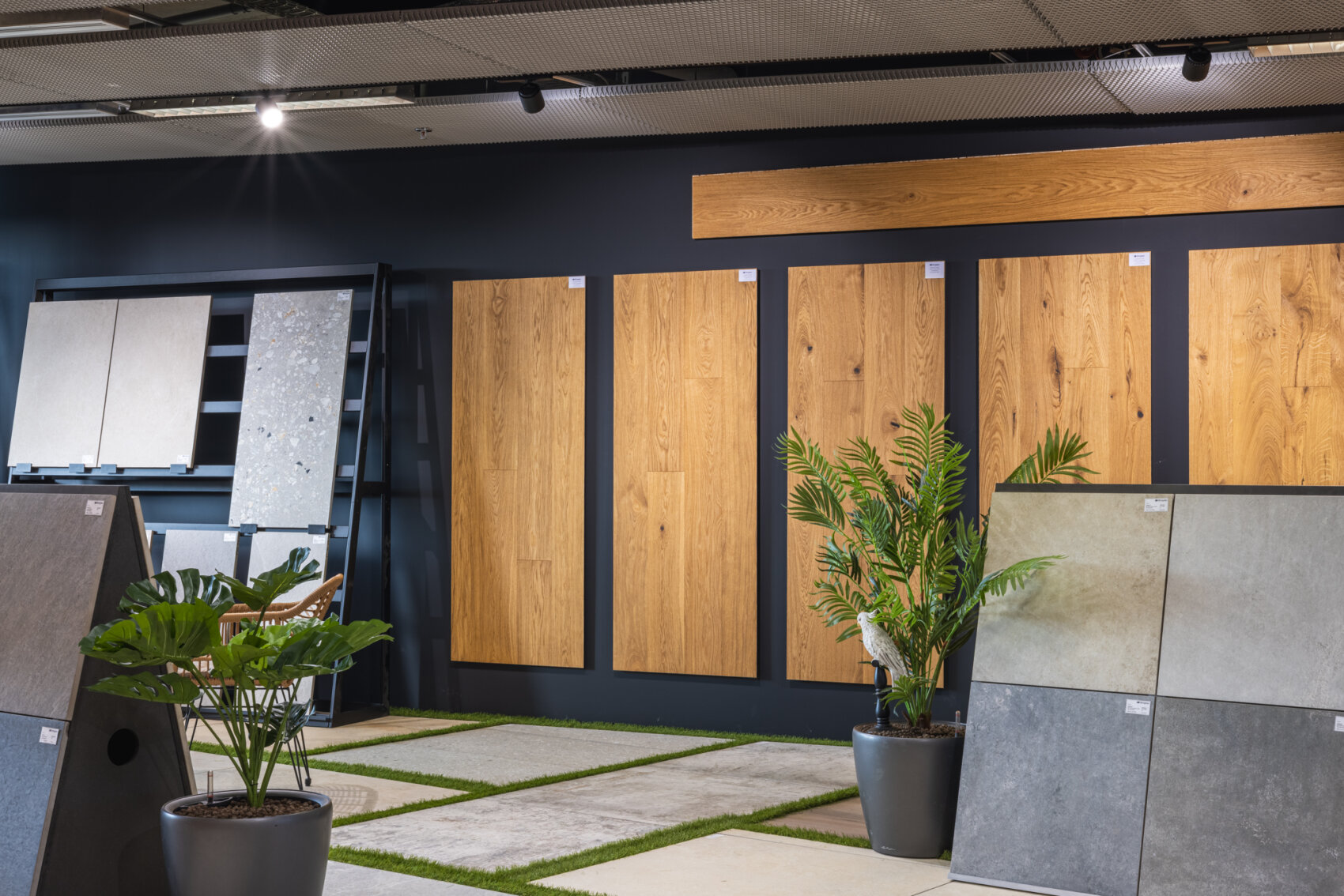 In einem Ausstellungsraum sind Muster von Holzpaneelen an einer dunklen Wand ausgestellt, während verschiedene Stein- und Fliesenmuster auf Regalen und dem Boden angeordnet sind. Zwei Topfpflanzen beleben den modernen, gut beleuchteten Raum.