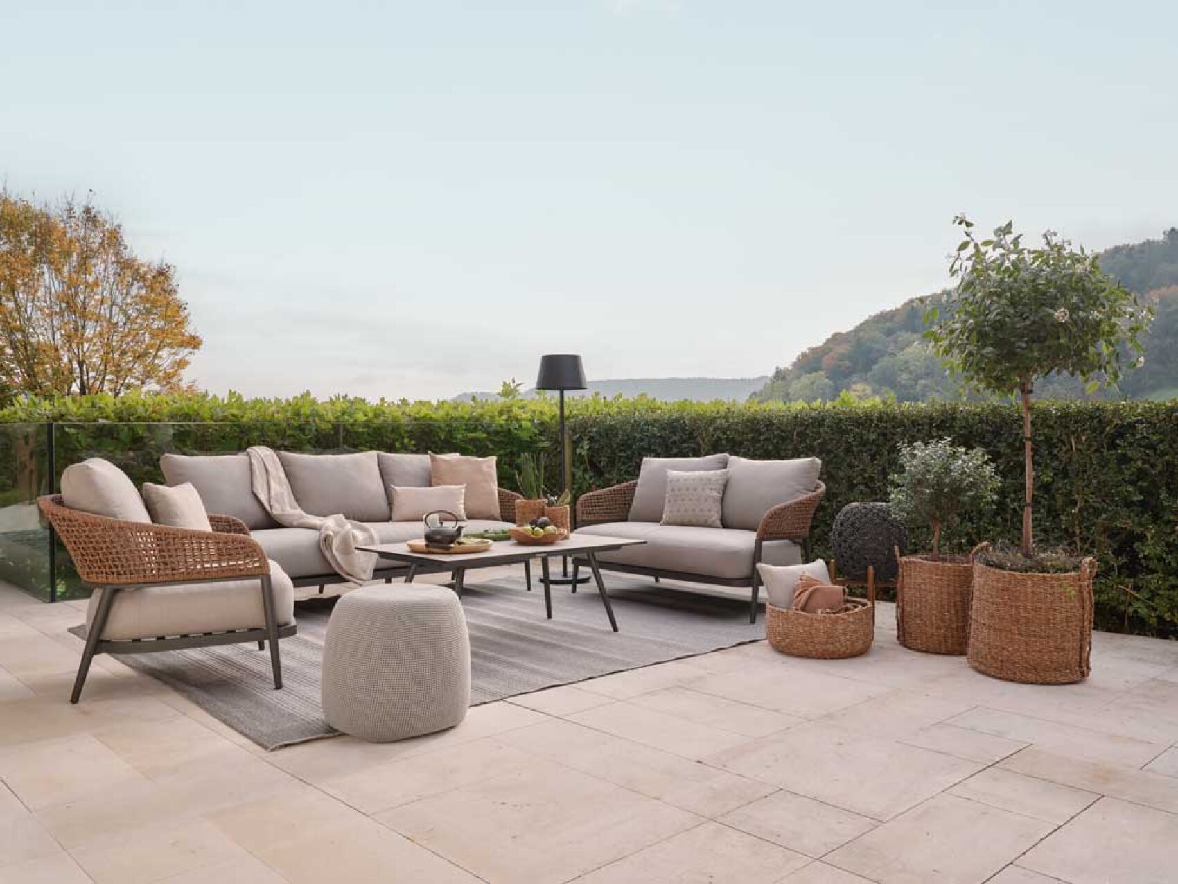 Terrasse im Freien mit einem Korbsofa und Sesseln mit beigen Kissen um einen Couchtisch, dekorativen Körben, Topfpflanzen, einem Hocker und einer Stehlampe, mit Blick ins Grüne und auf ferne Hügel unter einem klaren Himmel.