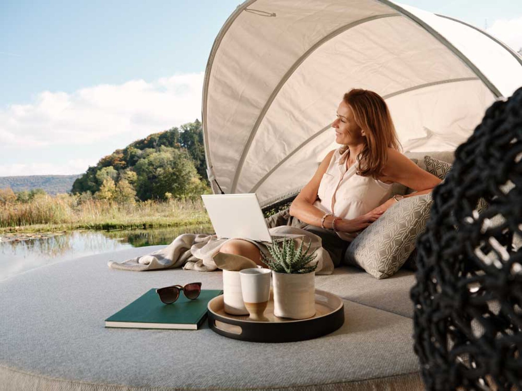 Eine Frau entspannt sich auf einer Outdoor-Lounge am Wasser und arbeitet an einem Laptop. Sie ist umgeben von Kissen, einem Tablett mit Tassen, einer kleinen Pflanze, einer Sonnenbrille und einem Notizbuch. Im Hintergrund sind Bäume und Hügel unter einem teilweise bewölkten Himmel zu sehen.
